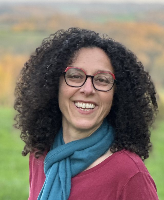 Portrait of Monique Shefer, smiling, in a teal scarf with autumn fields behind